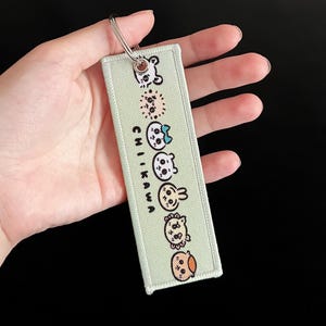 May include: A light green rectangular keychain with the word "Chikawa" printed vertically. The keychain features several cartoon animal characters. A silver metal ring is attached to the top.