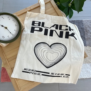 May include: A white canvas tote bag with black text that reads "BLACKPINK" and a stylized heart design. The names "JISOO", "ROSE", "JENNIE", and "LISA" are printed below the heart.