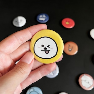 Op de afbeelding: Een gele ronde knoop met een cartoongezicht, vastgehouden in een hand. Het personage heeft een wit gezicht, zwarte ogen en een uitgestoken tong. Andere knopen met verschillende ontwerpen zijn zichtbaar.