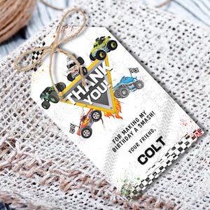 以下が含まれることがあります： 白いギフトタグ、モンスター・トラックをテーマにしたデザイン。カラフルなモンスター・トラックのイラスト、"THANK YOU!"の文字、"COLT"の名前が描かれています。タグは紐で結ばれています。