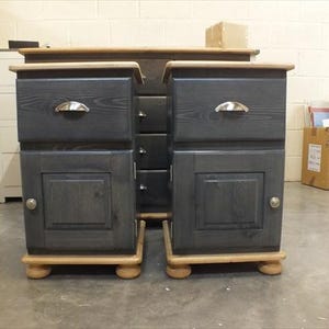 May include: Two black wooden bedside tables with a drawer and a door. The tables have a light wood top and bottom and are sitting on a concrete floor.
