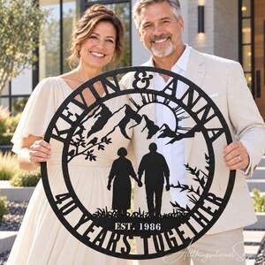 May include: A circular black metal wall sign featuring a mountain landscape, a sun, and a silhouette of two people holding hands. The text reads KEVIN & ANNA, 40 YEARS TOGETHER, and EST. 1986. The design includes trees and grass elements.