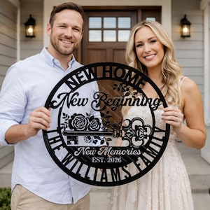 May include: A black metal sign with the words "NEW HOME, New Beginning, New Memories, THE WILLIAMS FAMILY, EST. 2026." The sign features a key and floral design. The sign is held by a couple in front of a house.