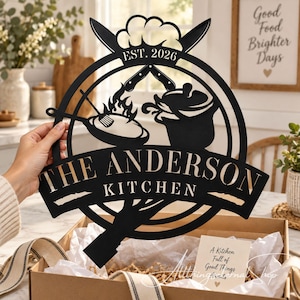 May include: A black metal circular wall sign featuring a cutout design of a frying pan with flames, a cooking pot, and crossed knives. The text reads EST. 2026, THE ANDERSON KITCHEN. The sign is held up against a blurred kitchen background.