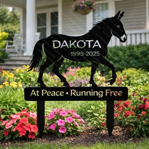 May include: Black metal garden stake featuring a silhouette of a donkey with the name "DAKOTA" and the years "1998-2025". The stake also includes the text "At Peace • Running Free". The stake is placed in a garden with colorful flowers.