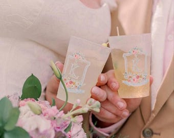 Peut inclure: Deux gobelets en plastique transparent ornés d'un écusson blanc avec la lettre K et un motif floral de fleurs roses et bleues. Les gobelets sont tenus ensemble et contiennent un liquide jaune clair avec un petit agitateur en bois visible.