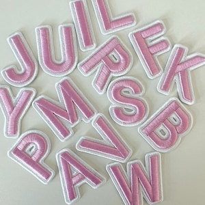 Könnte beinhalten: Rosa gestickte Buchstaben mit weißen Rändern. Die Buchstaben sind in verschiedenen Schriftarten und Größen, darunter J, U, L, R, F, K, Y, M, S, B, P, A, V und W. Die Buchstaben sind auf einer hellen Oberfläche verstreut.