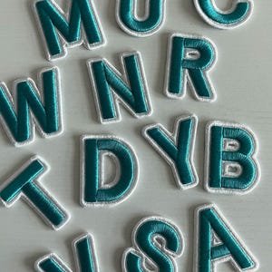 Puede incluir: Un conjunto de letras de fieltro de color verde azulado y blanco, que incluyen las letras M, U, C, W, N, R, T, D, Y, B, I, S y A. Las letras están bordeadas de blanco y tienen un centro verde azulado.