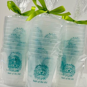 May include: Clear plastic cups with a teal dog illustration and the text "hair of the dog" are bundled in cellophane and tied with a green ribbon. The cups are stacked in sets, perfect for a party or event.
