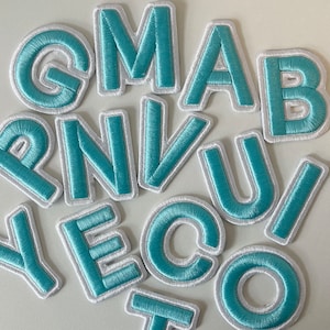 Pode incluir: Um conjunto de letras bordadas em tecido azul claro com uma borda branca. As letras, em uma fonte sem serifa, estão dispostas aleatoriamente. Provavelmente são patches ou apliques para roupas ou artesanato.