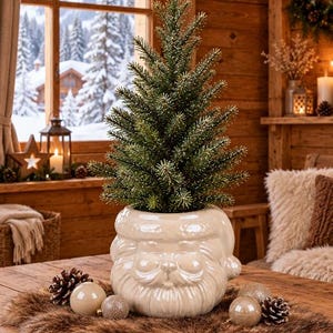 Könnte beinhalten: Ein kleiner künstlicher immergrüner Baum mit bereiften Spitzen in einem glänzenden cremefarbenen Keramikübertopf in Form eines Weihnachtsmann-Gesichts mit modelliertem Bart, Schnurrbart und Mützenrand auf einem braunen Kunstfell-Tischläufer.