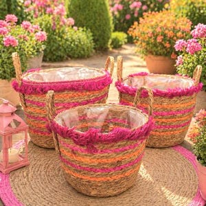 Könnte beinhalten: Drei gewebte Körbe mit orangefarbenen und rosa Streifen und Fransen, mit Griffen, stehen auf einem runden Jute-Teppich mit rosa Rand. Links steht eine rosa Laterne. Der Hintergrund zeigt einen Garten mit Blumen.