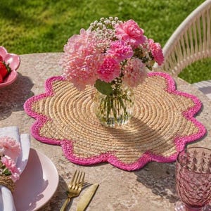 Könnte beinhalten: Eine Tischdekoration mit einem gewebten Platzset mit rosa gewelltem Rand, einer Vase mit rosa Blumen und einem rosa Teller mit Serviette und Blume. Eine Schale mit Erdbeeren und ein rosa Glas sind ebenfalls vorhanden.