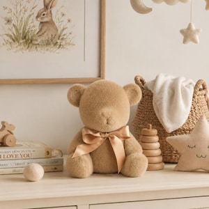 Könnte beinhalten: Ein beigefarbener Plüschteddybär mit einem glatten Satinband um den Hals. Der Bär hat eine weiche, flauschige Textur und sitzt aufrecht. Er ist umgeben von verschiedenen Kinderzimmerartikeln wie Büchern, einem geflochtenen Korb und einem Sternkissen.