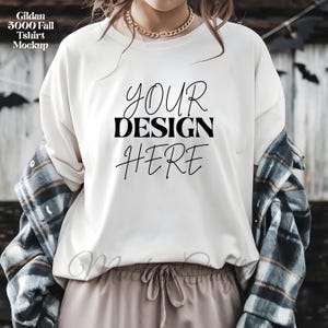 Può includere: Mockup di maglietta Gildan 5000 Fall color crema con la scritta "YOUR DESIGN HERE" in corsivo nero. Una camicia a quadri è drappeggiata sulle spalle. La modella indossa una collana a catena dorata e pantaloni beige.