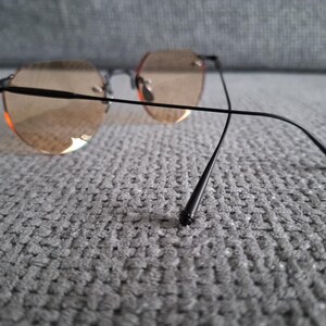 May include: Rimless sunglasses with round, light brown tinted lenses and black metal frames. The temples extend back with a slight curve. The glasses are resting on a gray, textured surface.