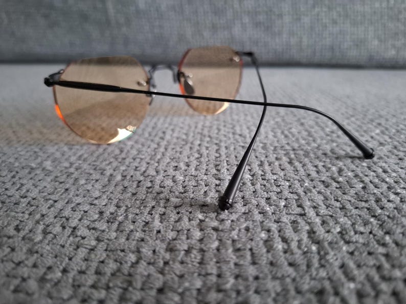 May include: Rimless sunglasses with round, light brown tinted lenses and black metal frames. The temples extend back with a slight curve. The glasses are resting on a gray, textured surface.