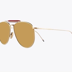 May include: Gold-tone aviator sunglasses with brown lenses. The sunglasses feature a double bridge, a red accent on the top bar, and thin temples. The lenses are a warm brown color, and the frame has a shiny finish.