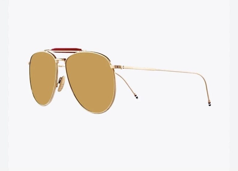 May include: Gold-tone aviator sunglasses with brown lenses. The sunglasses feature a double bridge, a red accent on the top bar, and thin temples. The lenses are a warm brown color, and the frame has a shiny finish.