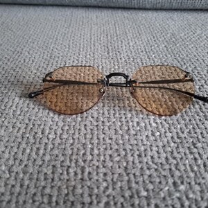 May include: Rimless sunglasses with brown tinted lenses and a black bridge and arms. The lenses are a rounded hexagonal shape. The sunglasses are resting on a gray textured surface.