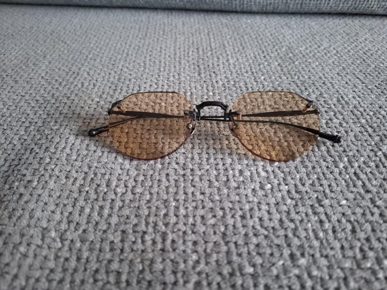 May include: Rimless sunglasses with brown tinted lenses and a black bridge and arms. The lenses are a rounded hexagonal shape. The sunglasses are resting on a gray textured surface.