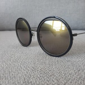 May include: Round sunglasses with black frames and reflective lenses. The lenses have a gold tint, and the frames are thick and circular. The temples are thin and dark, and the sunglasses are resting on a light gray surface.