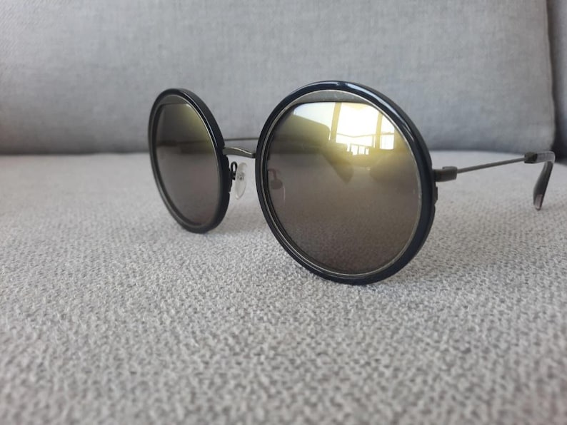 May include: Round sunglasses with black frames and reflective lenses. The lenses have a gold tint, and the frames are thick and circular. The temples are thin and dark, and the sunglasses are resting on a light gray surface.