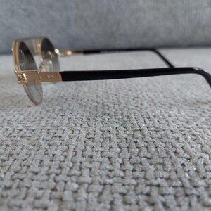 Puede incluir: Gafas con montura dorada y patillas negras. Las lentes redondas est&aacute;n engastadas en una montura dorada, con varillas negras que se extienden hasta la oreja. Las gafas descansan sobre una superficie texturizada de color gris claro.