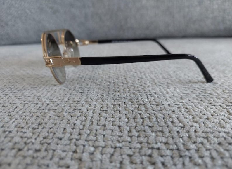 Puede incluir: Gafas con montura dorada y patillas negras. Las lentes redondas est&aacute;n engastadas en una montura dorada, con varillas negras que se extienden hasta la oreja. Las gafas descansan sobre una superficie texturizada de color gris claro.