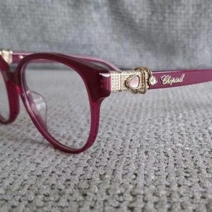 May include: Deep magenta eyeglasses with clear lenses and a rounded frame shape. The temples feature gold-tone metal accents with a small heart-shaped detail, embedded crystals, and the Chopard brand name printed in gold lettering on the side.
