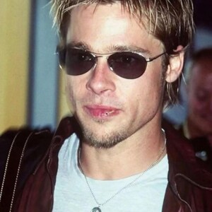 May include: A person wearing oval-shaped sunglasses with a silver frame. The person is wearing a light gray t-shirt, a silver chain necklace with a pendant, and a burgundy leather jacket.