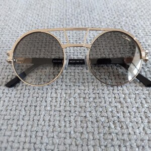 Puede incluir: Gafas de sol Cazal doradas con lentes redondas y un dise&ntilde;o de doble puente. Las lentes son de color gris oscuro y las patillas son negras. Las gafas de sol tienen un aspecto moderno y elegante.