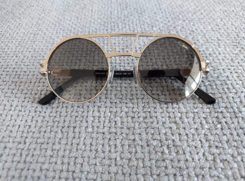 Puede incluir: Gafas de sol Cazal doradas con lentes redondas y un dise&ntilde;o de doble puente. Las lentes son de color gris oscuro y las patillas son negras. Las gafas de sol tienen un aspecto moderno y elegante.