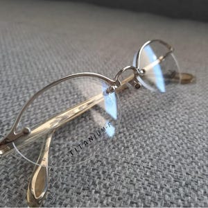 Puede incluir: Gafas de titanio doradas con montura redonda sin montura y lentes transparentes. El texto "TITANIUM-F" está impreso en la lente derecha.