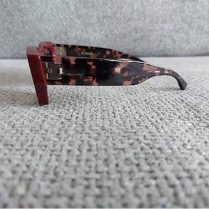 May include: Red and tortoiseshell sunglasses with a thick frame. The sunglasses have a rectangular shape and a dark lens.