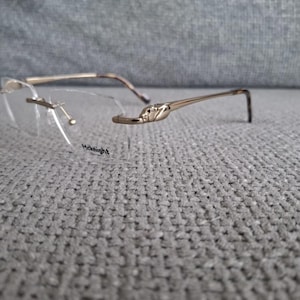 May include: Rimless spectacles with gold-coloured frames. The lenses are clear, and the temples have a decorative design. The text "Midnight" is printed on the lens. The glasses are resting on a grey textured surface.