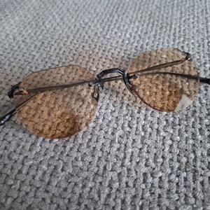 May include: Rimless sunglasses with brown tinted lenses and a black bridge and arms. The lenses are a rounded, octagonal shape. The glasses are resting on a gray, textured surface.