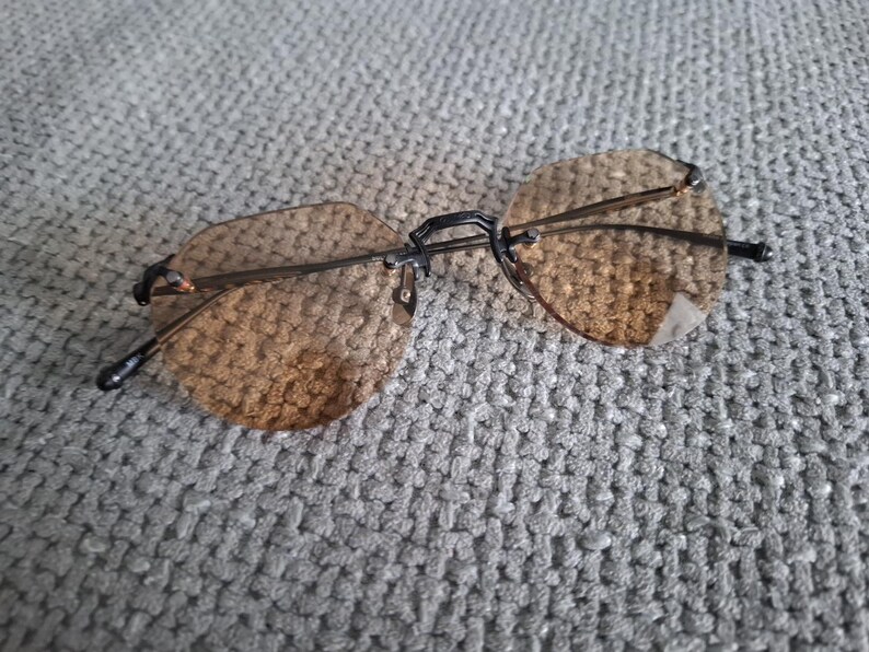 May include: Rimless sunglasses with brown tinted lenses and a black bridge and arms. The lenses are a rounded, octagonal shape. The glasses are resting on a gray, textured surface.