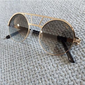 Puede incluir: Gafas de sol doradas con lentes redondas y un dise&ntilde;o de puente &uacute;nico. Las lentes son de color gris degradado y las monturas tienen una est&eacute;tica geom&eacute;trica moderna. Las patillas son negras.