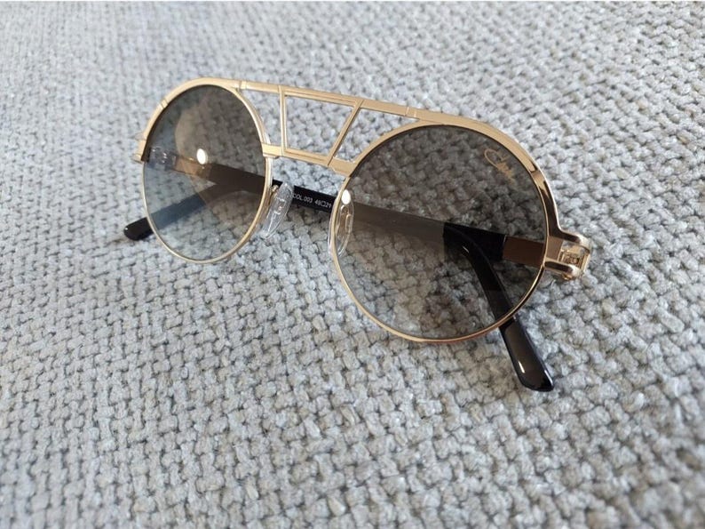 Puede incluir: Gafas de sol doradas con lentes redondas y un dise&ntilde;o de puente &uacute;nico. Las lentes son de color gris degradado y las monturas tienen una est&eacute;tica geom&eacute;trica moderna. Las patillas son negras.