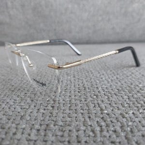 Op de afbeelding: Bril zonder montuur met heldere glazen en goudkleurige accenten. De veren hebben een gestructureerd zilveren patroon en zwarte uiteinden. De merknaam "Midnight" is op het glas gedrukt.