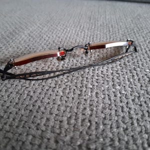 May include: Rimless eyeglasses with a brown and silver frame and black temples. The glasses have a rectangular shape and are resting on a gray textured surface. The temples have a decorative design.