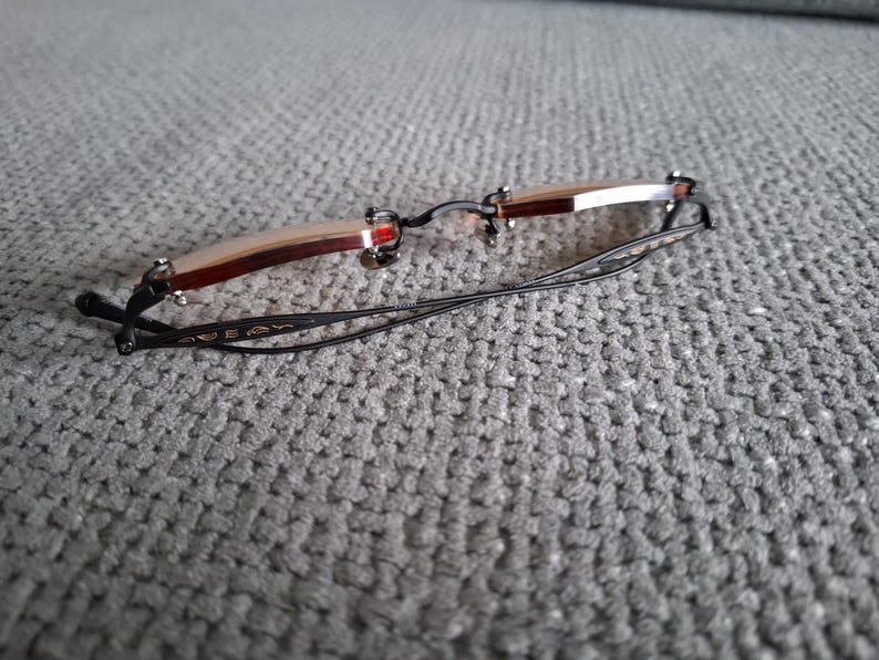 May include: Rimless eyeglasses with a brown and silver frame and black temples. The glasses have a rectangular shape and are resting on a gray textured surface. The temples have a decorative design.