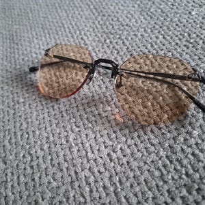 May include: Rimless sunglasses with brown tinted lenses and a black metal frame. The lenses have a geometric shape. The glasses are resting on a gray, textured surface.