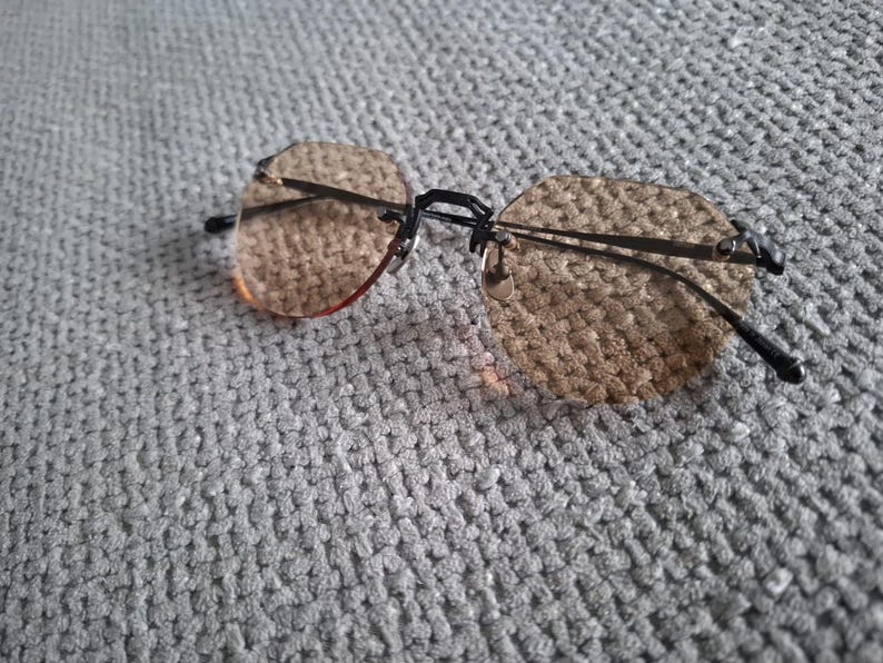 May include: Rimless sunglasses with brown tinted lenses and a black metal frame. The lenses have a geometric shape. The glasses are resting on a gray, textured surface.