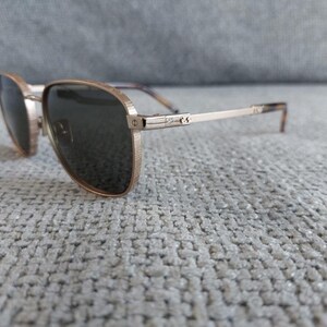 Puede incluir: Gafas de sol de aviador doradas con lentes oscuras. Las monturas tienen un acabado texturizado y las patillas son finas con un patr&oacute;n sutil cerca de las patillas. Las gafas de sol descansan sobre una superficie gris texturizada.