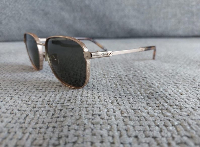 Puede incluir: Gafas de sol de aviador doradas con lentes oscuras. Las monturas tienen un acabado texturizado y las patillas son finas con un patr&oacute;n sutil cerca de las patillas. Las gafas de sol descansan sobre una superficie gris texturizada.
