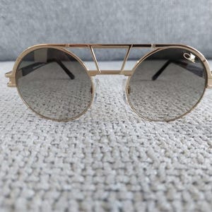 Puede incluir: Gafas de sol doradas con lentes redondas y un dise&ntilde;o de doble puente. Las gafas tienen una montura dorada y lentes oscuras. El dise&ntilde;o incluye una barra superior y un peque&ntilde;o logotipo en el lateral. Accesorio de moda.