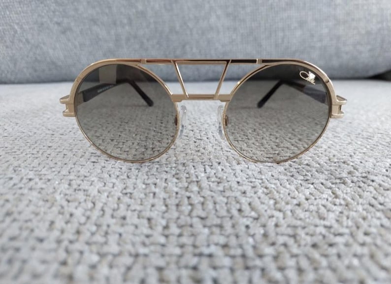 Puede incluir: Gafas de sol doradas con lentes redondas y un dise&ntilde;o de doble puente. Las gafas tienen una montura dorada y lentes oscuras. El dise&ntilde;o incluye una barra superior y un peque&ntilde;o logotipo en el lateral. Accesorio de moda.