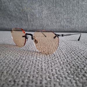May include: Rimless sunglasses with light brown tinted lenses. The glasses have a black frame and temples, with a small gold-colored accent at the nose bridge. The lenses are a unique octagonal shape.